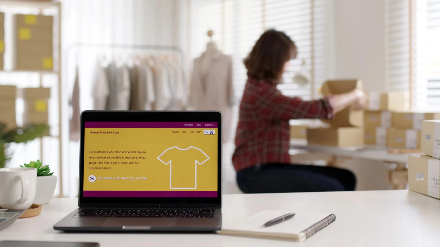 Laptop que muestra una tienda online y de fondo una mujer empacando.
